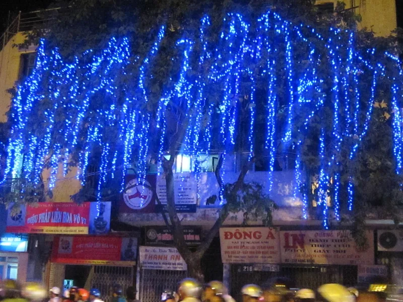 Blue string lights drape a tree over a shopfront street in Hanoi at night, with motorbike riders passing below.