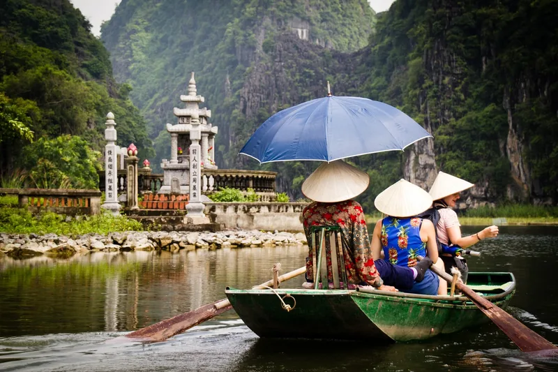 Boatwomen in conical hats row tourists past a temple gate along a river surrounded by karst mountains in Tam Coc, Ninh Binh.