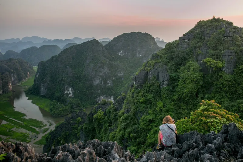 A traveler sits atop a rocky viewpoint overlooking the karst mountains and winding river of Ninh Binh at dusk.