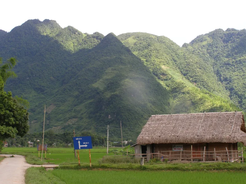 A thatched-roof stilt house sits beside rice paddies below jagged green mountains in Mai Chau valley.