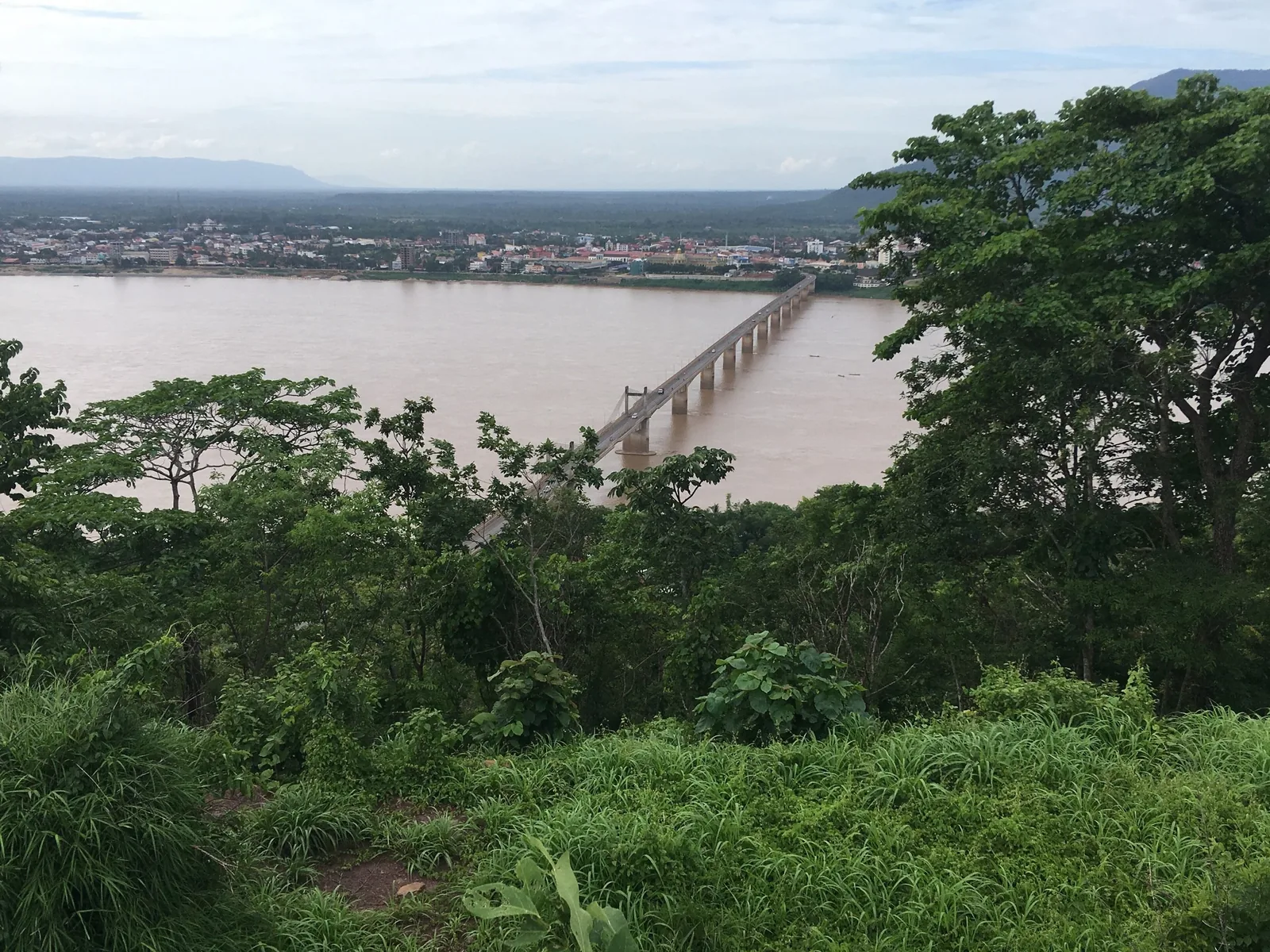 A long bridge spans the muddy Mekong River near Pakse, seen through green foliage from a hillside viewpoint.