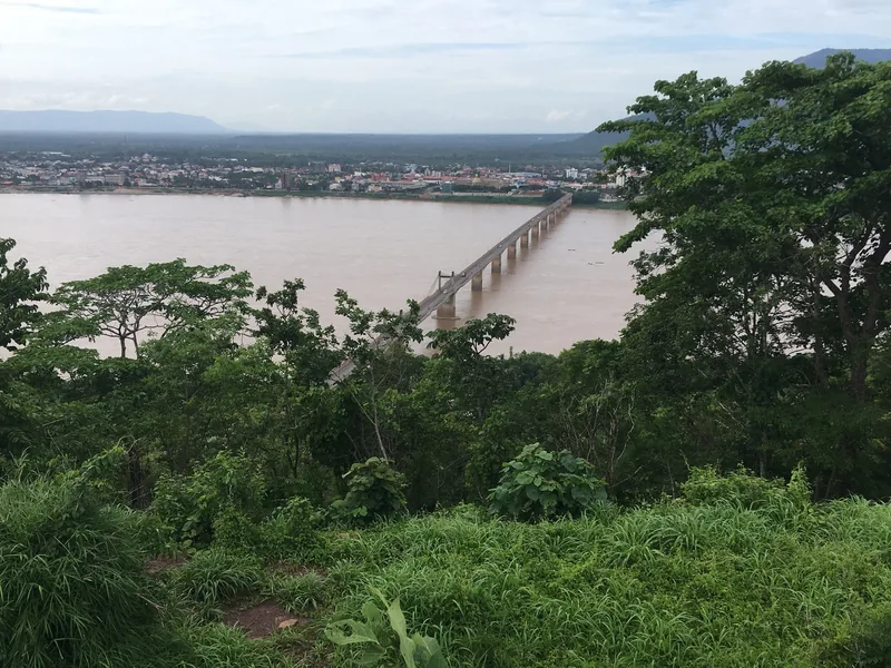 A long bridge spans the muddy Mekong River near Pakse, seen through green foliage from a hillside viewpoint.