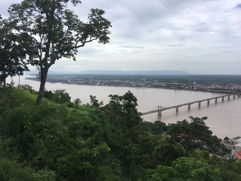 A hillside view overlooks the wide Mekong River, a bridge, and the town of Pakse under a cloudy sky.