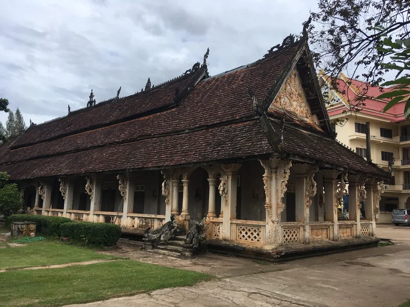 A weathered tiered-roof temple with ornate carved columns stands at Wat Luang in Pakse, Laos.