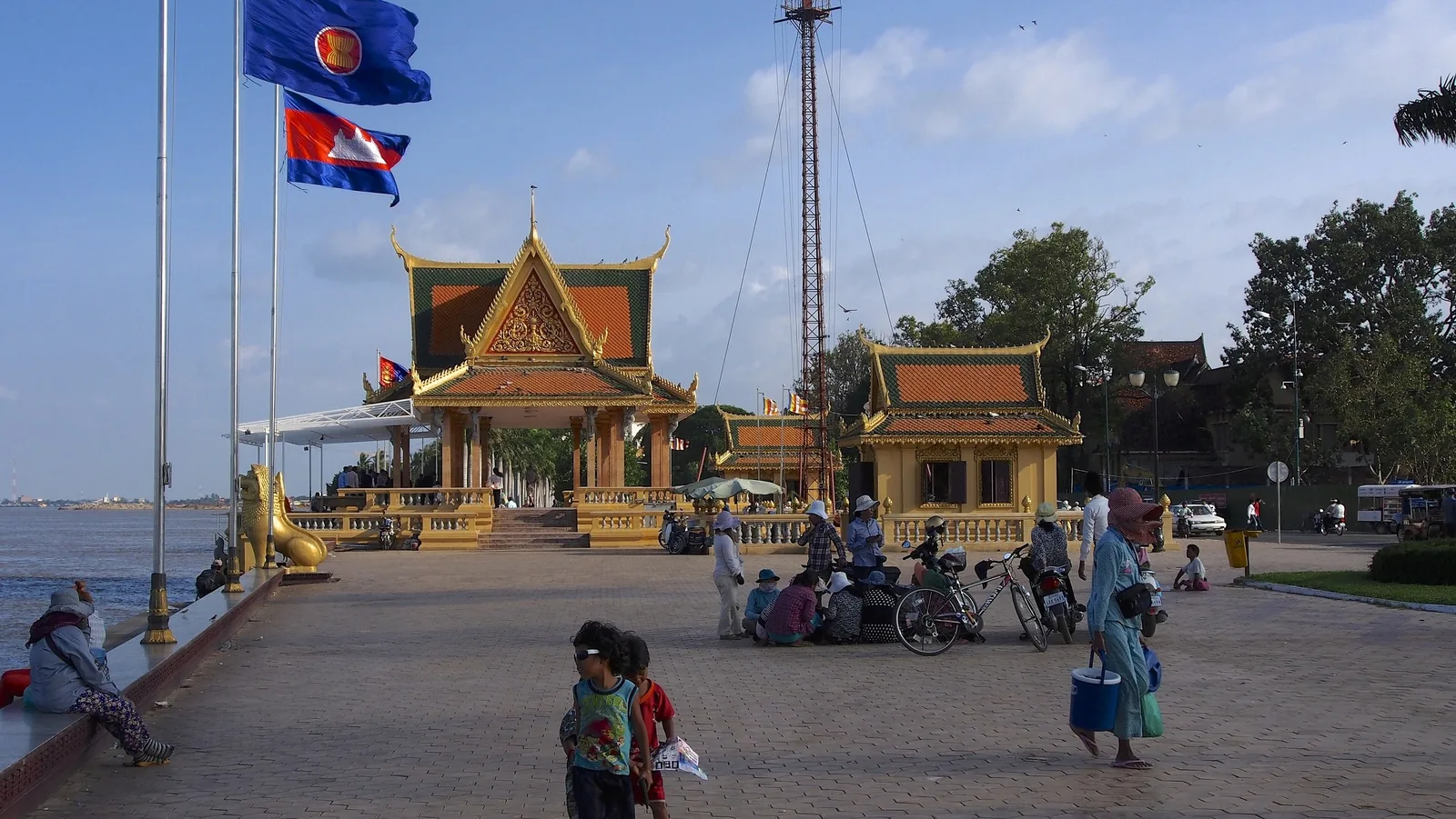 Golden pavilions and flags line the riverside promenade in Phnom Penh, with locals walking by.