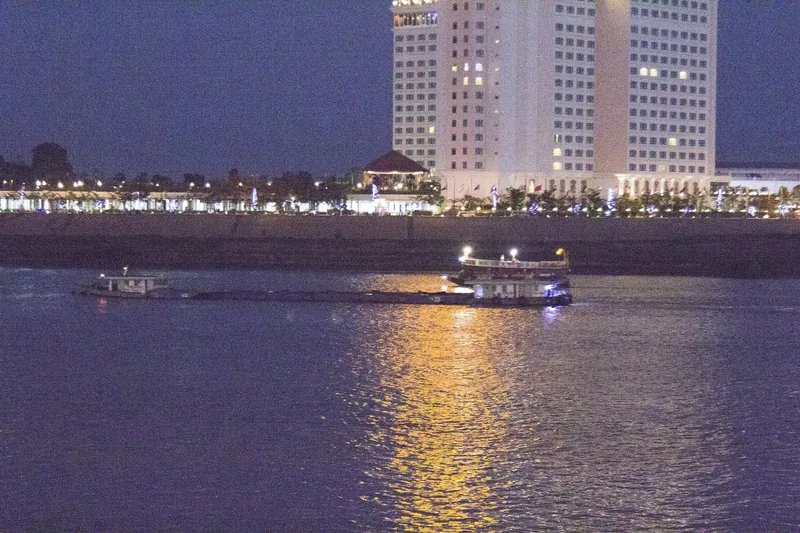 A cargo boat glides along the Tonle Sap river at night beneath the illuminated Phnom Penh skyline.
