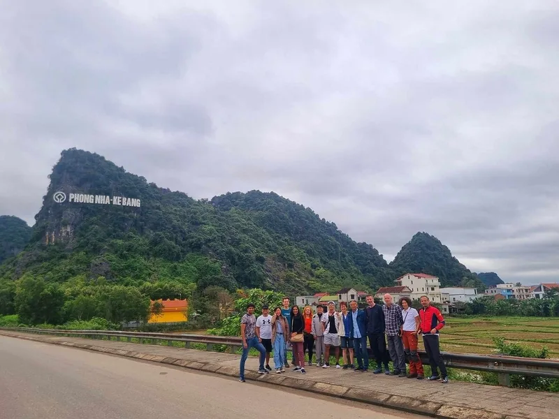 A group of travelers poses along a roadside with the Phong Nha-Ke Bang National Park sign on a limestone mountain behind.