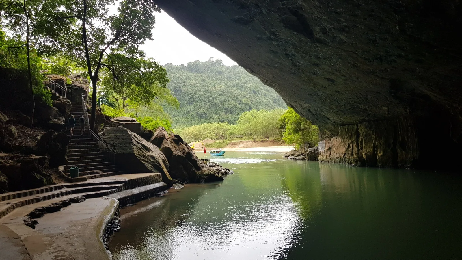 A stone stairway leads beside a calm river flowing beneath a cave entrance surrounded by jungle in Phong Nha.