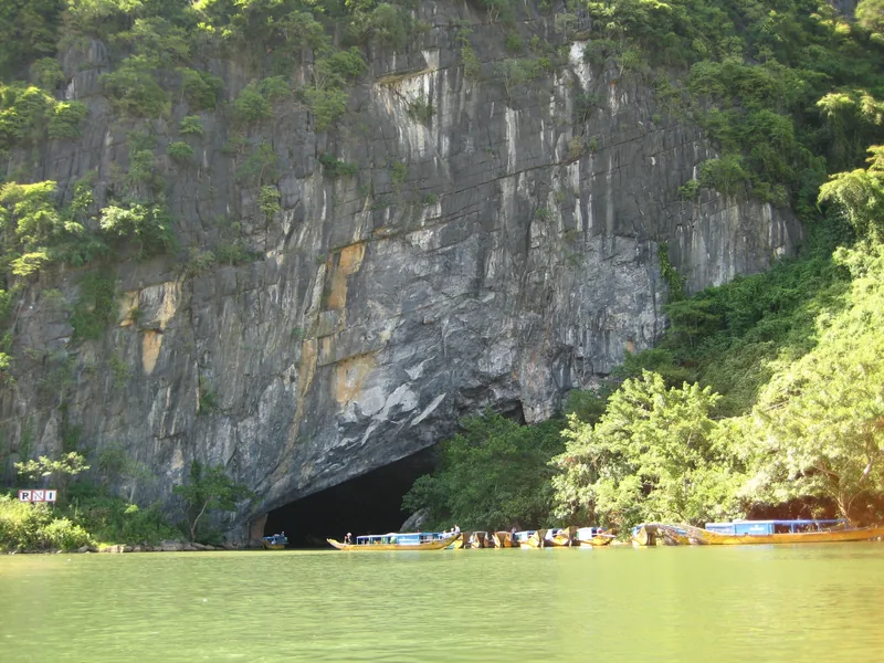 Wooden tourist boats gather at the entrance of Phong Nha cave beneath a towering limestone cliff.