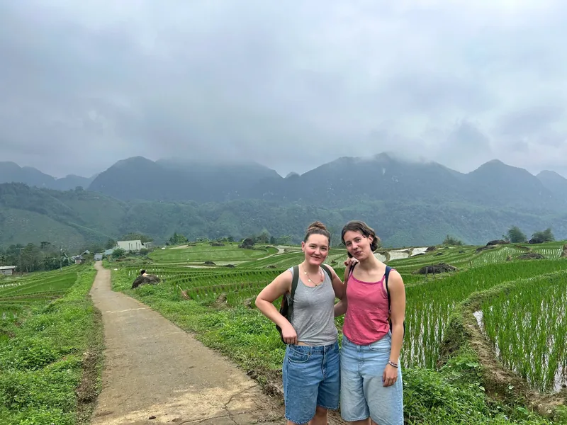 Two women pose on a path through green rice terraces with misty mountains behind them in Pu Luong, Vietnam.