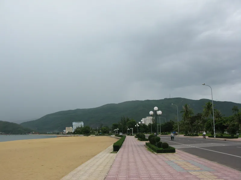 An empty beachfront promenade with a paved walkway and road runs along Quy Nhon's coast under overcast skies.