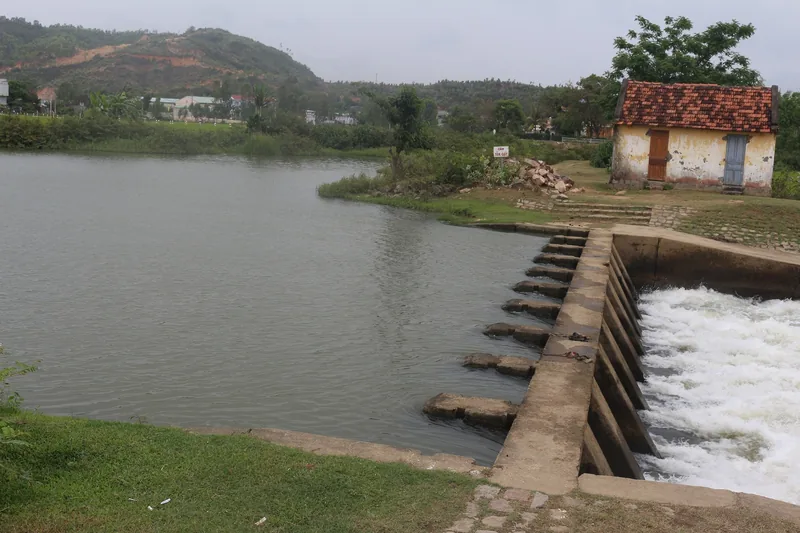 A concrete spillway releases water from a reservoir near a small tiled-roof house in the hills around Quy Nhon.