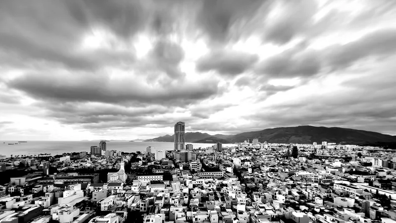 A black-and-white aerial view shows Quy Nhon's dense rooftops beneath dramatic clouds, with a tall tower and coastal.