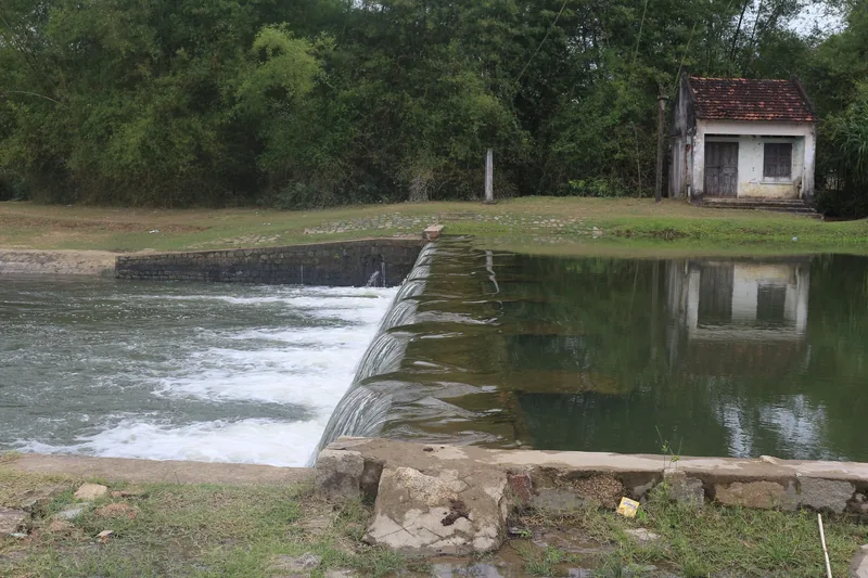 A small weir spills water into a calm river beside an old tiled-roof house near Quy Nhon.