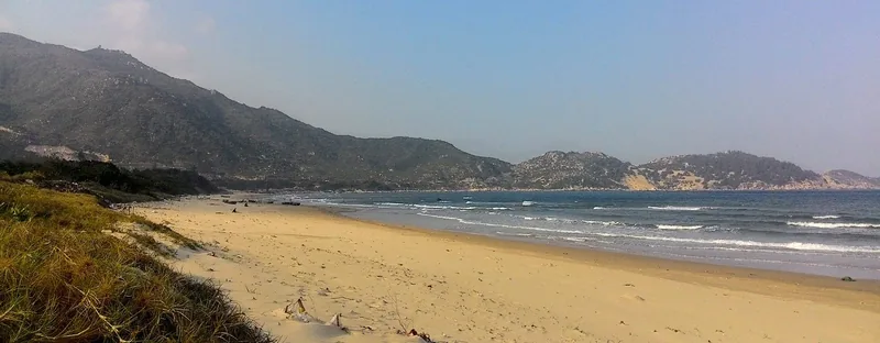 A wide sandy beach curves along the coastline near Phuong Mai Peninsula in Quy Nhon, with green hills rising in the distance.