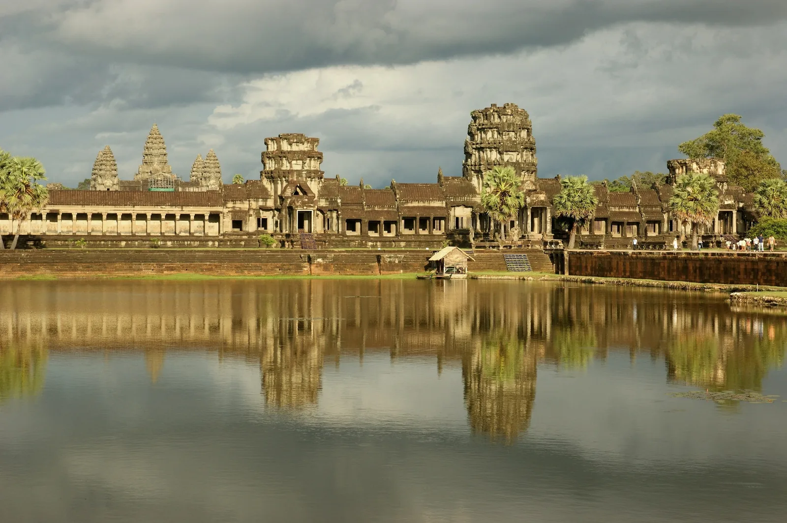 Angkor Wat's stone towers and galleries reflect in the surrounding moat under a cloudy sky in Siem Reap.