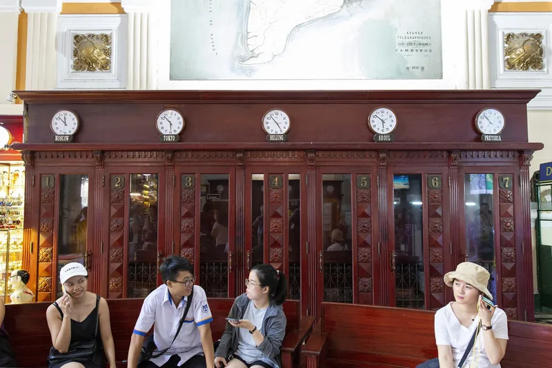 Visitors sit on a wooden bench below vintage world clocks and an old map inside Saigon Central Post Office in Ho Chi Minh.