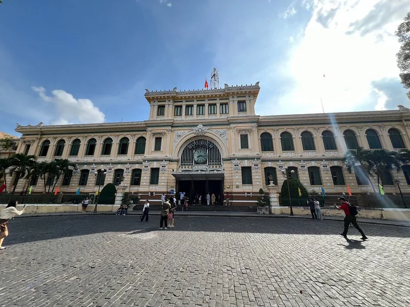 The colonial facade of Saigon Central Post Office rises above a cobbled plaza with visitors strolling by.