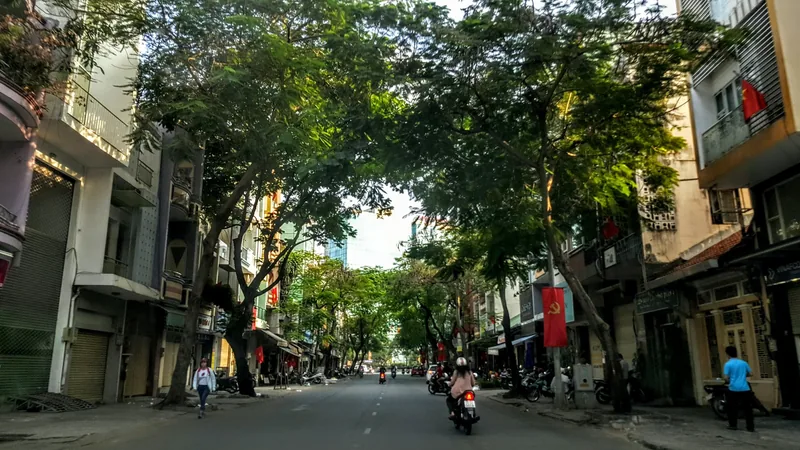 A tree-lined street in Ho Chi Minh City with motorbikes, shops, and flags under a canopy of green leaves.