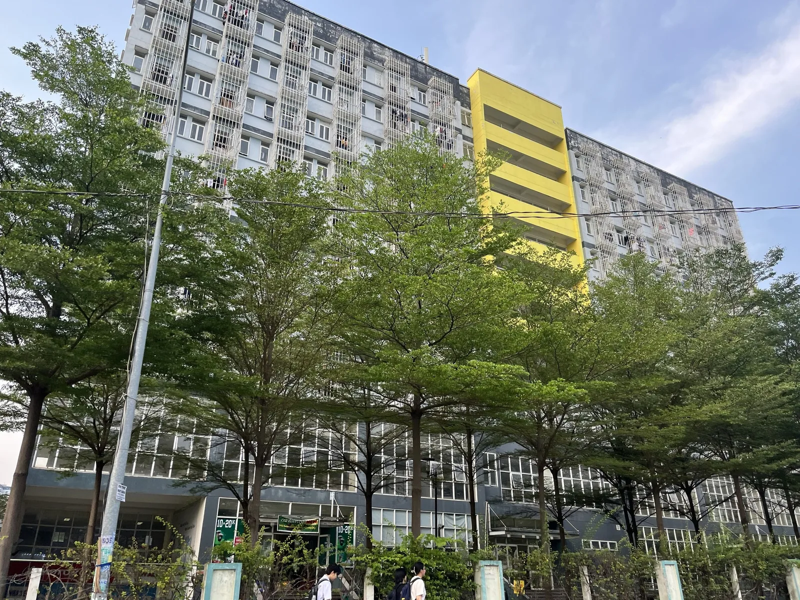 A tall apartment block with barred balconies and a bright yellow stairwell stands behind trees in Ho Chi Minh City.