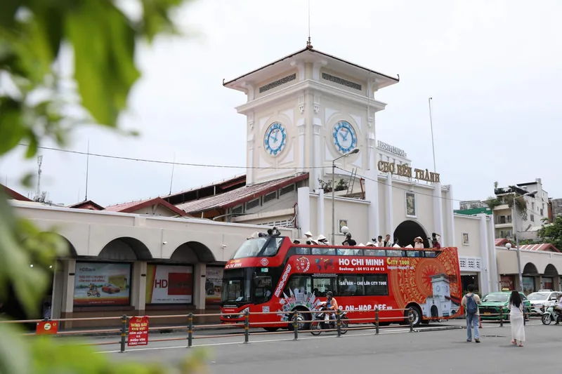 A red double-decker sightseeing bus passes the clock tower entrance of Ben Thanh Market in Ho Chi Minh City.