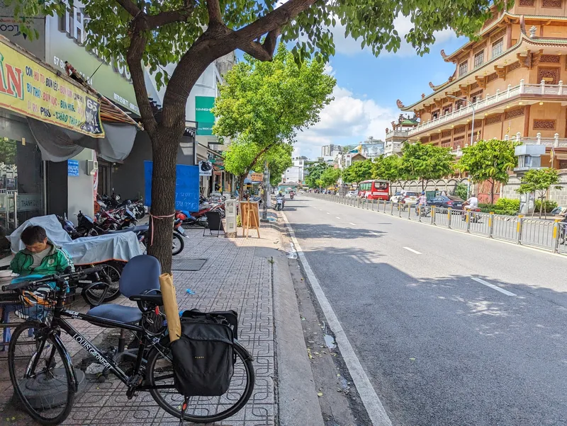 A tree-lined street in Ho Chi Minh City with shops, parked motorbikes and a bicycle near Giac Nguyen Temple.