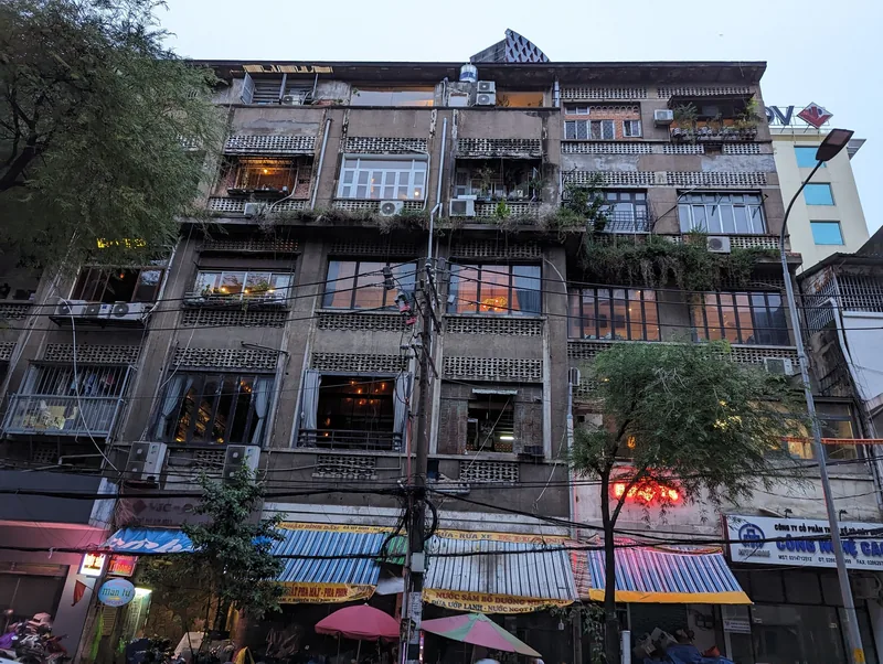 An old apartment block with balconies, plants, and tangled wires rises above shops in Ho Chi Minh City.