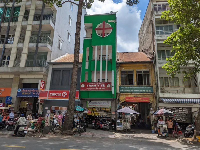 A tall green medical clinic building with a red cross sign stands among narrow shophouses on a busy Ho Chi Minh City street.