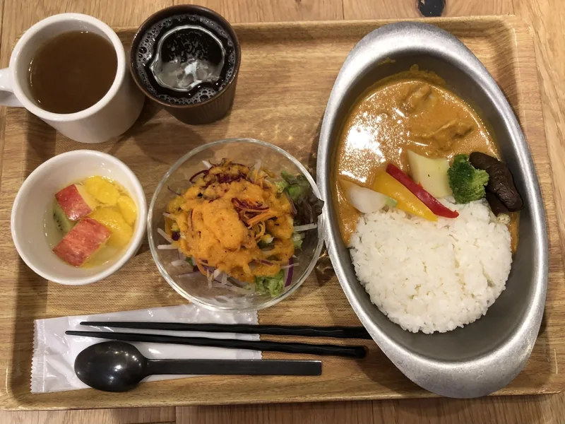 A tray meal with curry rice, salad, fruit cup, tea and iced coffee served on a wooden table.