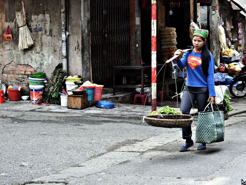 A vendor carries a shoulder pole basket of vegetables along a weathered street in Ho Chi Minh City.