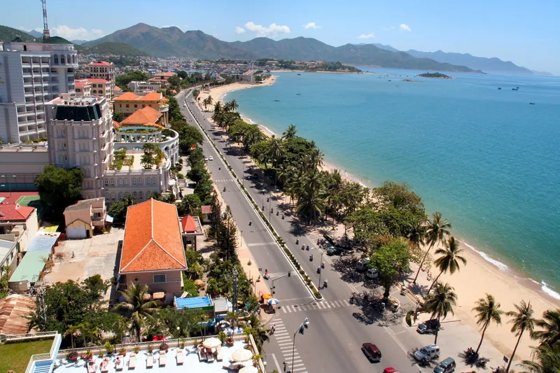 Aerial view of Nha Trang's coastal road lined with palm trees, hotels, and a sandy beach beside turquoise water.
