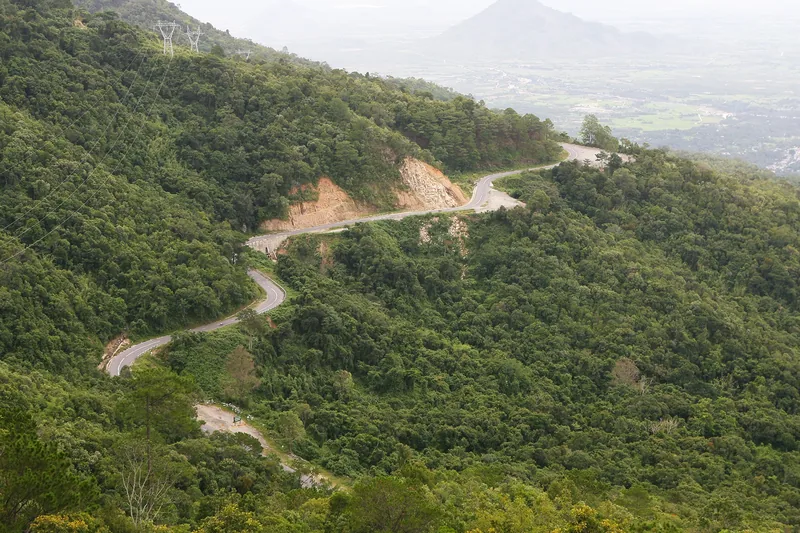 A winding mountain road cuts through dense forest with distant peaks near Da Lat, Lam Dong.