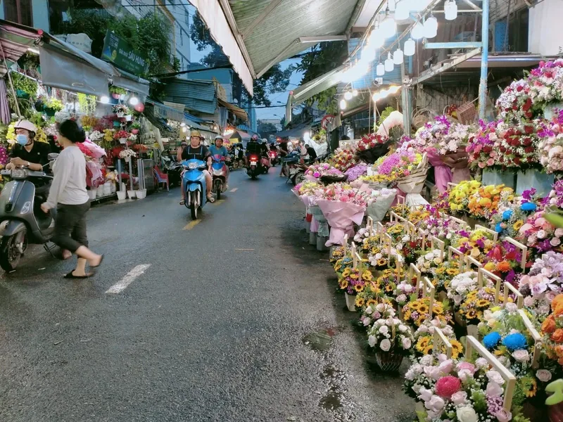 A colorful flower market street in Saigon lined with bouquets while motorbikes and pedestrians pass by at dusk.