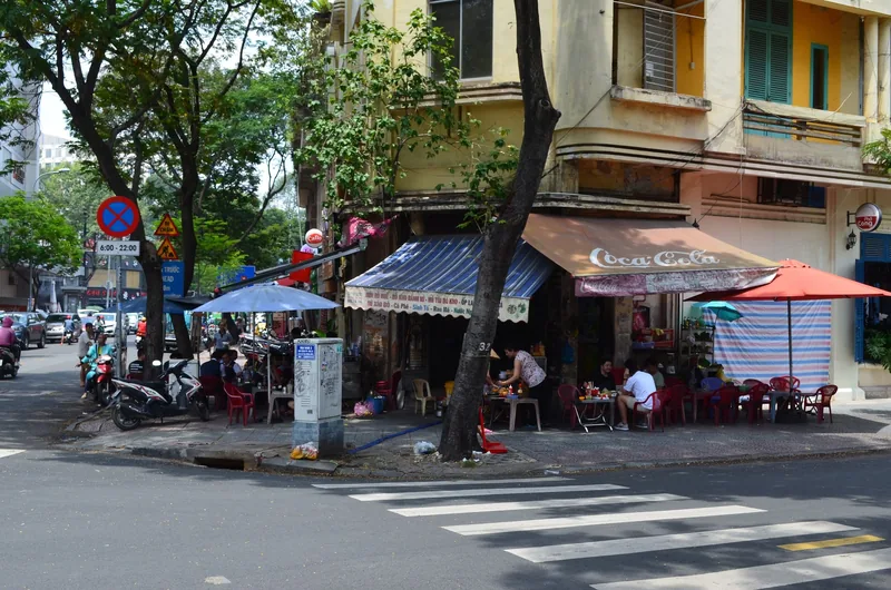 A street corner cafe with plastic chairs and colorful umbrellas sits beneath old apartment buildings in Ho Chi Minh City.