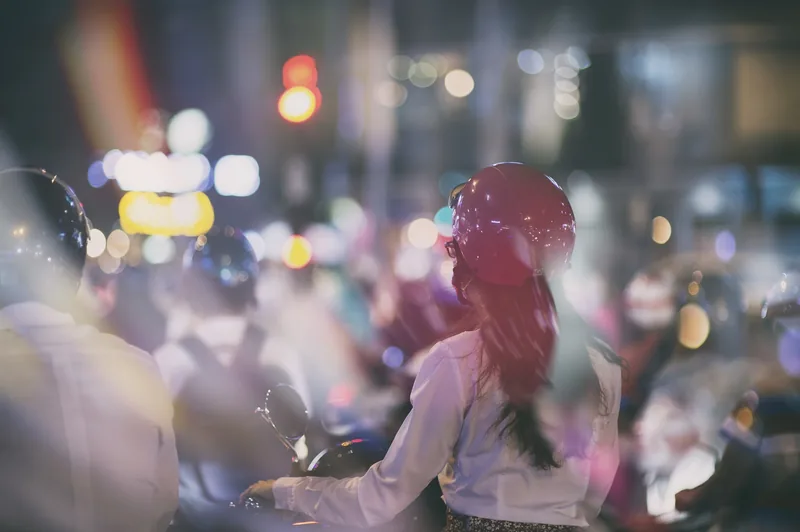 A woman on a motorbike wears a pink helmet amid a bustling, light-filled night traffic scene in Ho Chi Minh City.