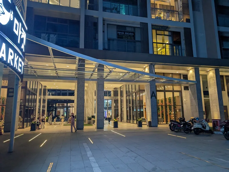 Modern apartment building entrance with glass canopy and lit storefronts at dusk in Ho Chi Minh City's Phu My Hung area.