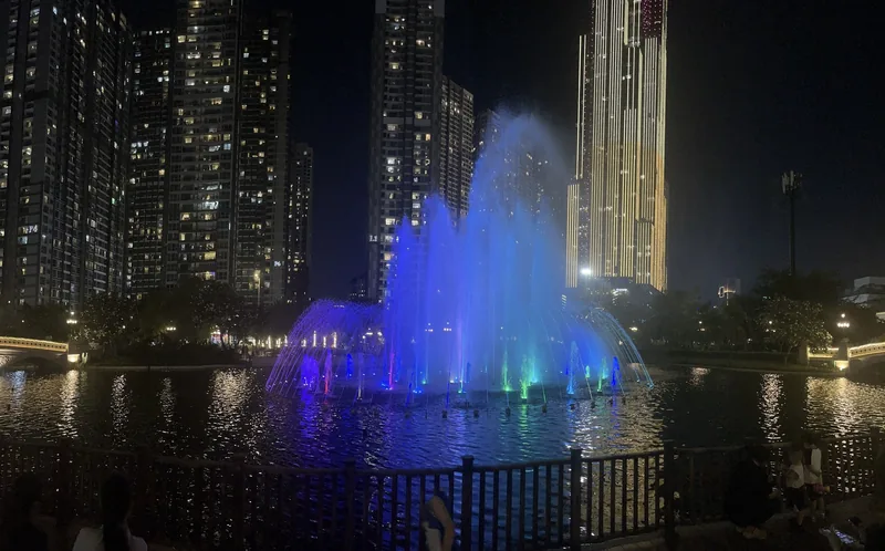 A blue-lit musical fountain glows on the lake at Vinhomes Central Park with high-rise towers behind it at night in Ho Chi.