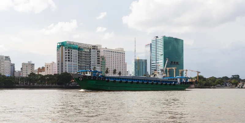 A green cargo barge cruises the Saigon River past the modern skyline of Ho Chi Minh City.