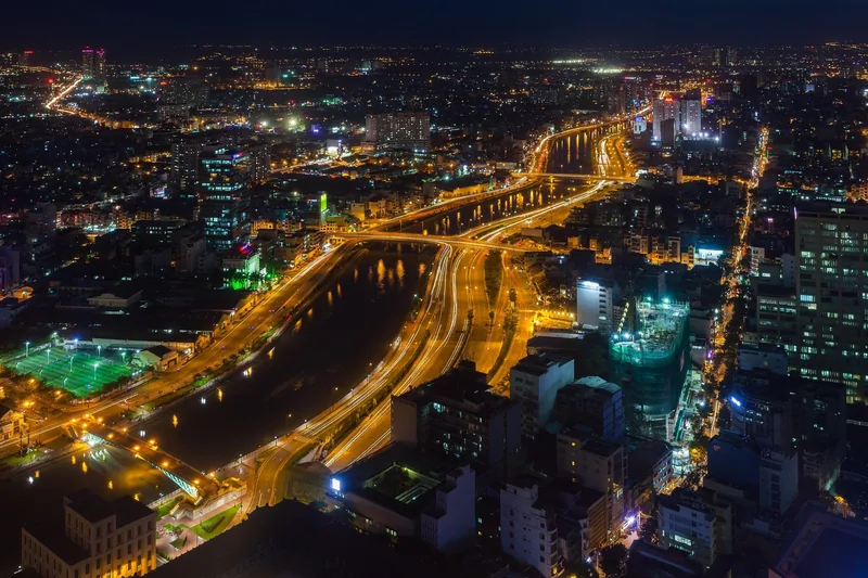 Aerial night view of Ho Chi Minh City with illuminated streets and a winding river cutting through dense buildings.