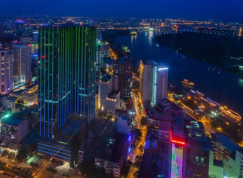 Aerial night view of Ho Chi Minh City skyscrapers glowing with neon lights along the Saigon River.