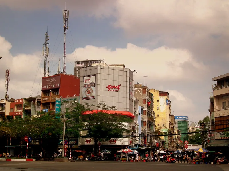 A busy street corner in Ho Chi Minh City lined with shops, signage, and antenna-topped buildings under a partly cloudy sky.