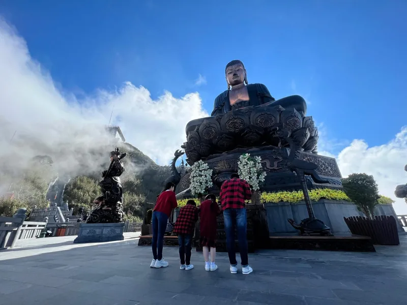 Visitors admire the giant bronze Buddha statue atop Fansipan mountain surrounded by clouds under a blue sky.