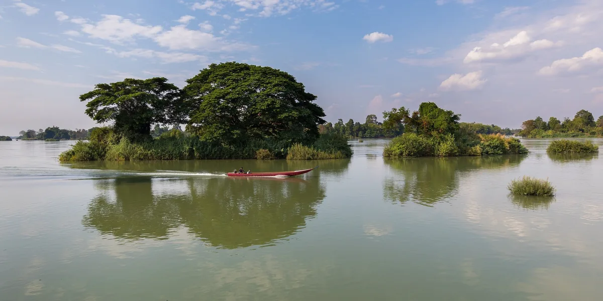 A long-tail boat glides past small wooded islets on the calm Mekong River in Si Phan Don, Laos.