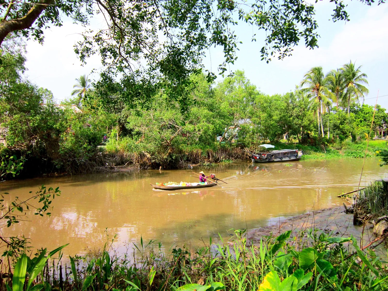 A small wooden boat carrying vendors paddles along a muddy canal lined with lush greenery near Can Tho.