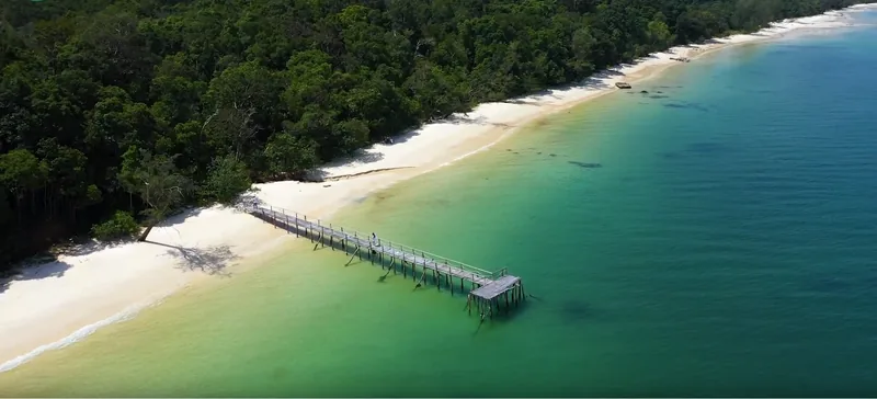 Aerial view of a wooden pier extending into turquoise water along a forested beach near Rach Vem, Phu Quoc.