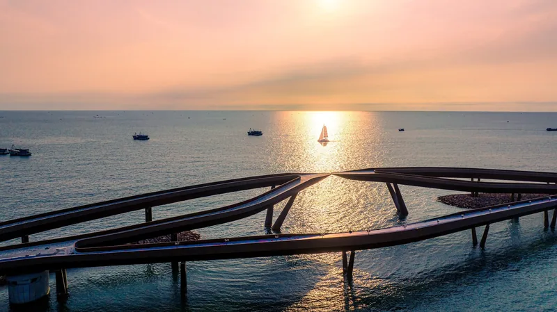 Aerial view of the curving Kiss Bridge over the sea at sunset in Phu Quoc, with a sailboat silhouetted on the horizon.