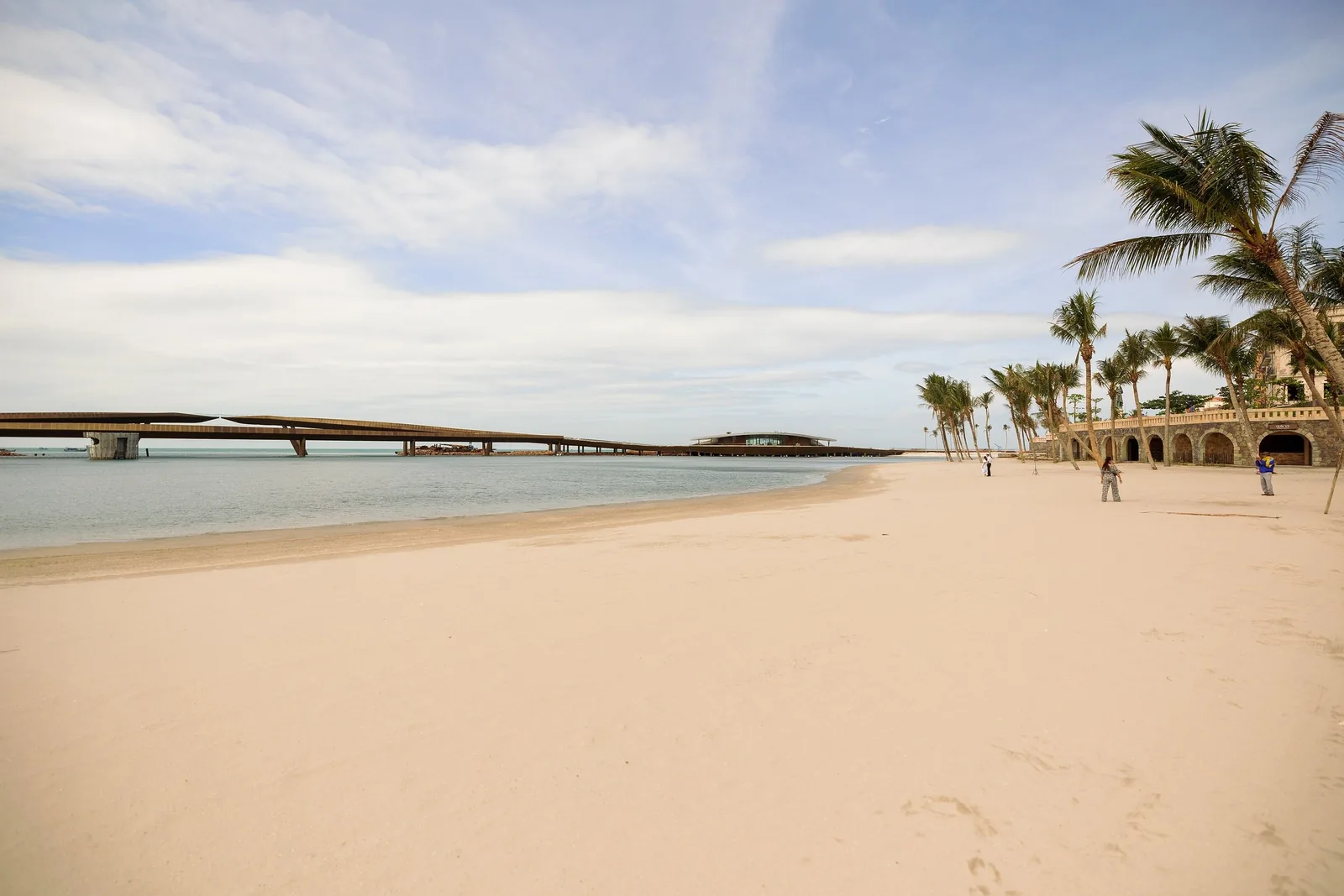 A long bridge stretches over calm water beside a sandy beach lined with palm trees in Phu Quoc.