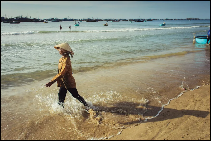 A woman in a conical hat walks through shallow surf near a fishing fleet at Mui Ne beach.