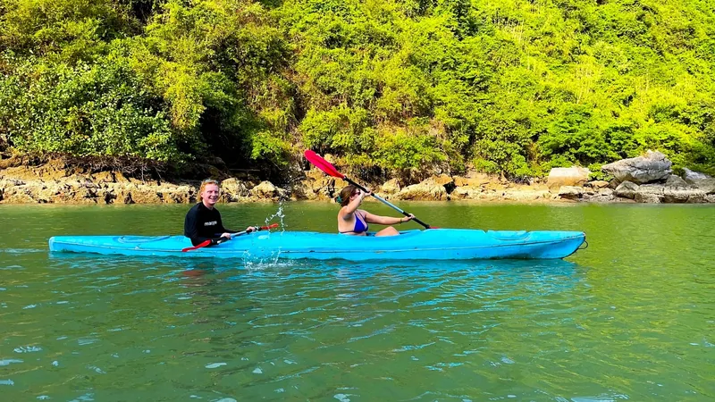 Two people paddle a blue kayak along a green lagoon lined with limestone cliffs in Lan Ha Bay.
