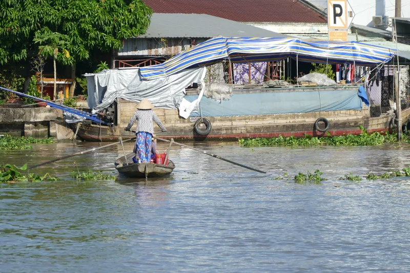 A person wearing a conical hat rows a small wooden boat toward a larger houseboat on a Mekong Delta river.
